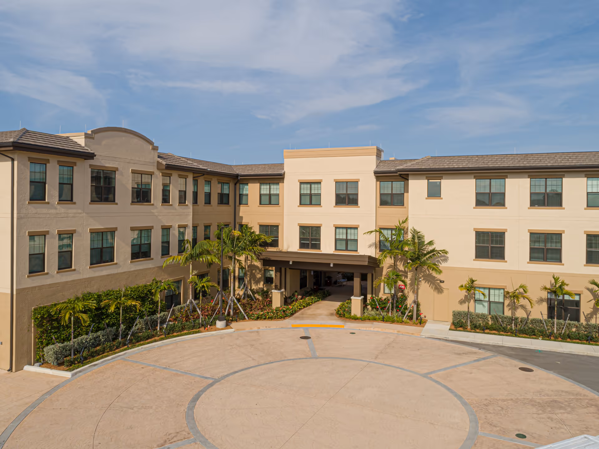 Exterior view of a three-story senior living facility building with beige and light brown walls, multiple windows, palm trees, and landscaped greenery around the entrance driveway under a partly cloudy sky.