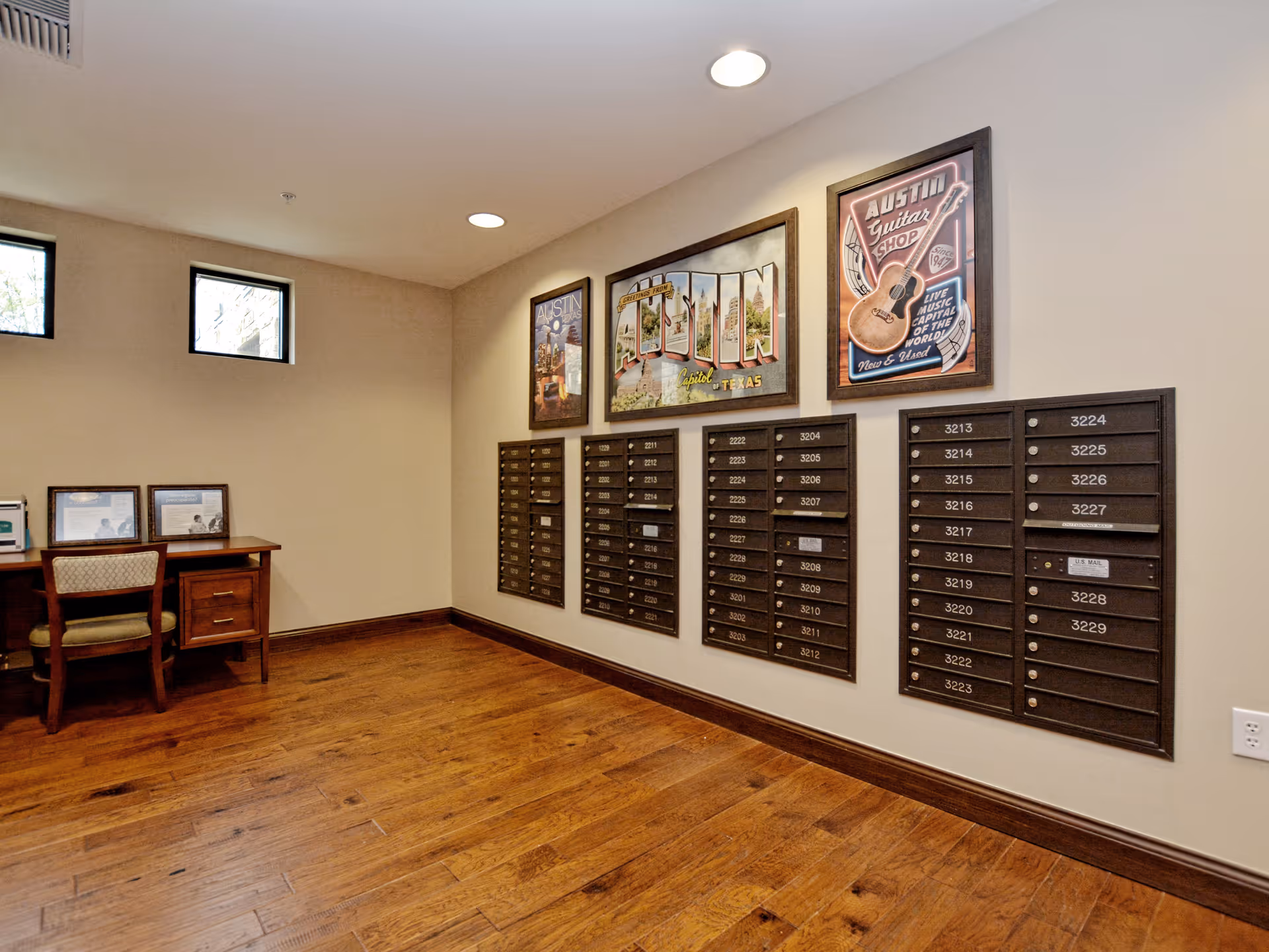 Interior mailroom with rows of wall-mounted apartment mailboxes, framed posters above, and a small desk and chair on hardwood floors.