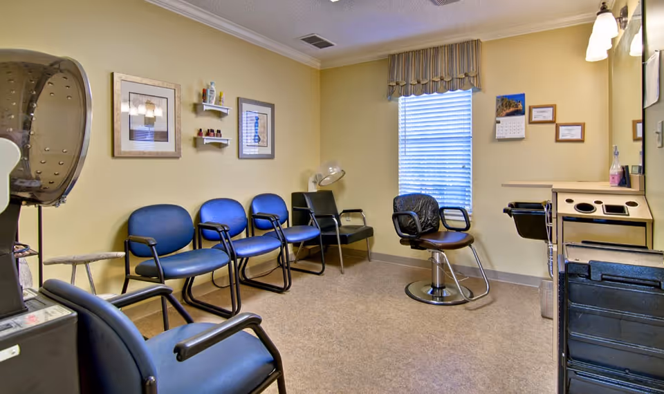 A small salon or beauty room with several blue chairs lined up against a yellow wall, a hair dryer hood on the left, a salon chair with a black cover in front of a window with blinds, and a counter with a mirror and hair care products on the right.