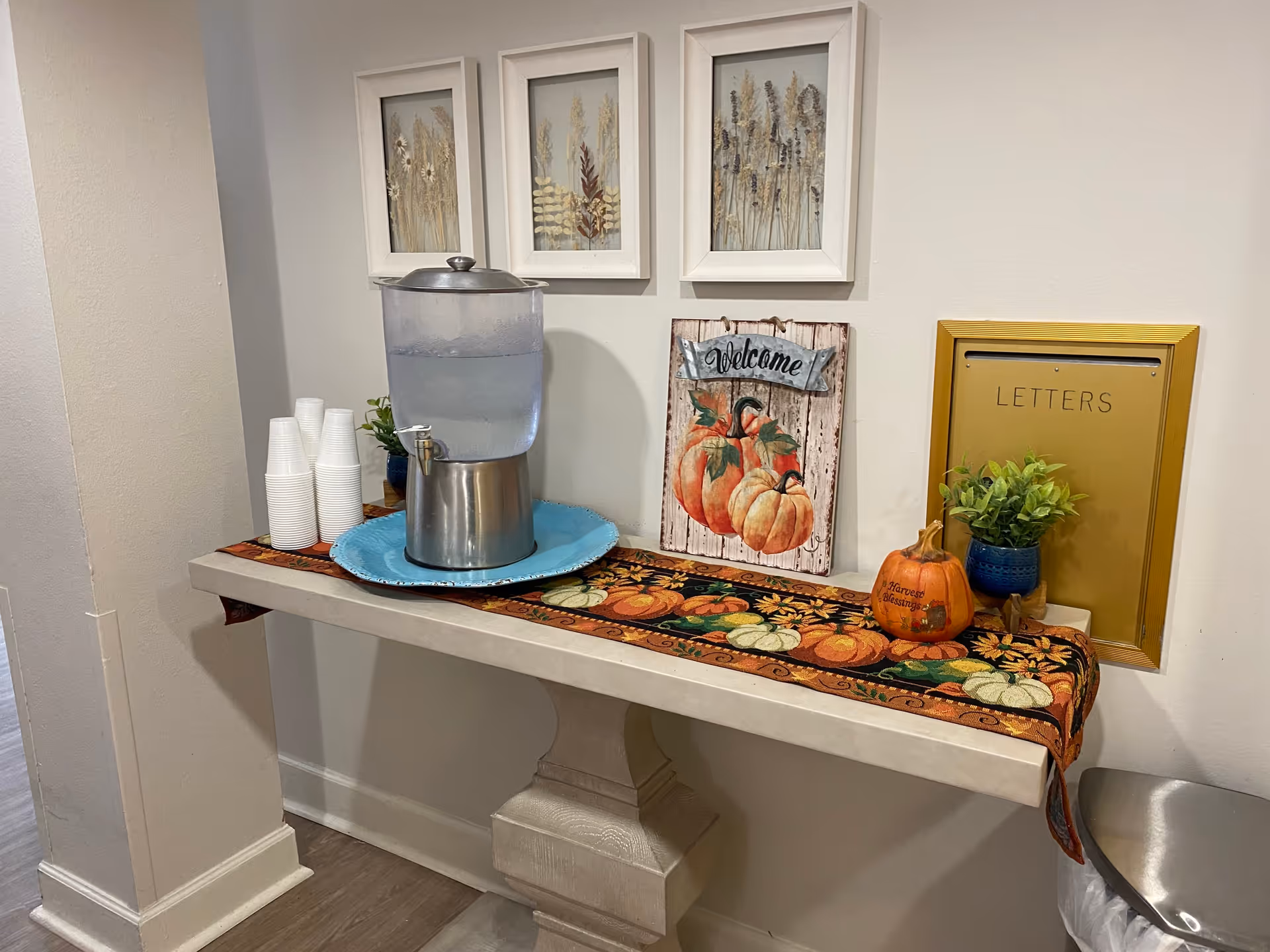 A small table against a wall decorated with a fall-themed runner featuring pumpkins. On the table is a water dispenser with a stack of white disposable cups, a small potted plant, a decorative sign with pumpkins and the word 'Welcome', a small pumpkin decoration with the words 'Harvest Blessings', and a gold-colored mail slot labeled 'LETTERS'. Above the table are three framed pictures of dried plants.