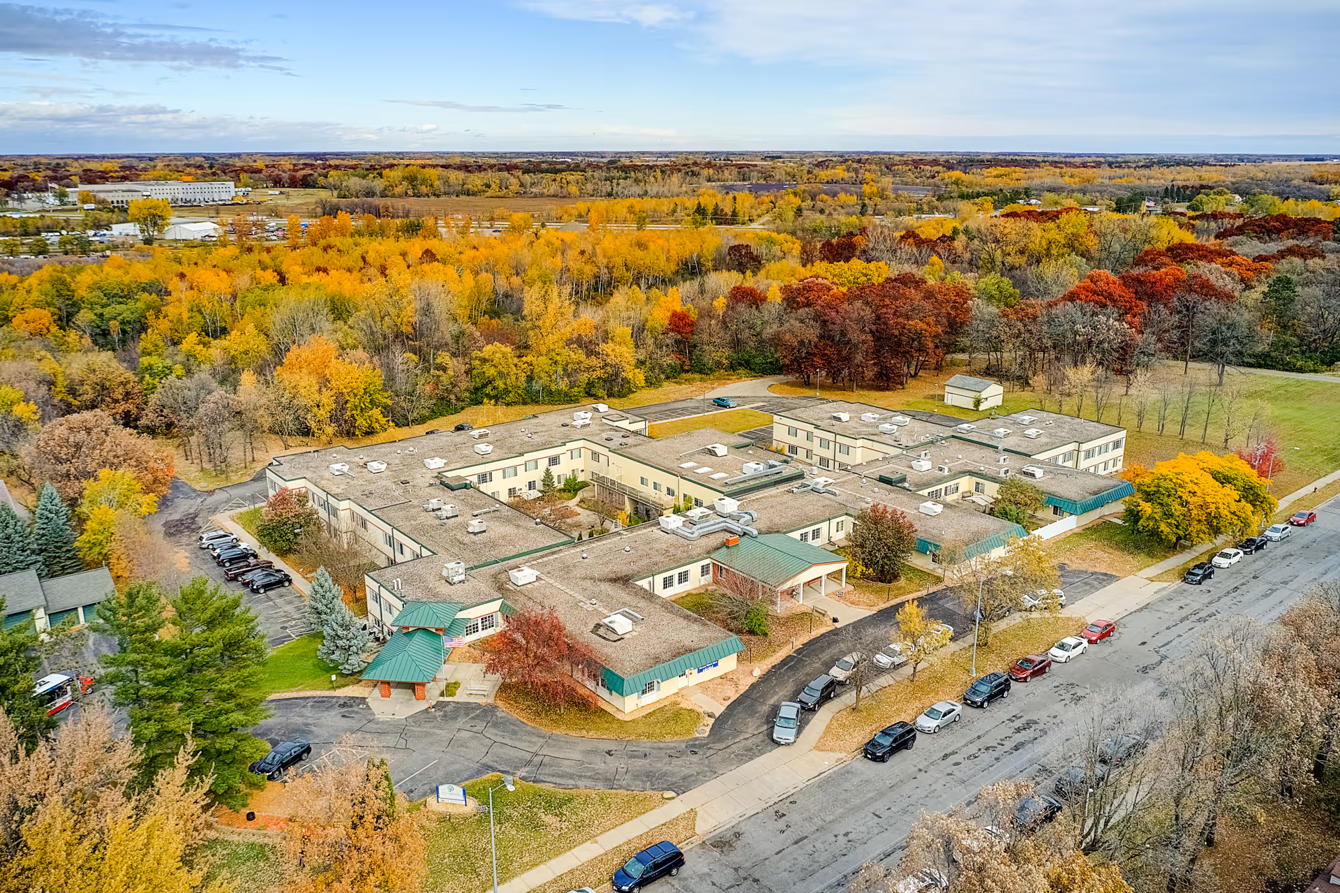 Aerial view of Nature's Point Assisted Living facility surrounded by trees with autumn foliage in shades of yellow, orange, and red. The building has a flat roof and green awnings over entrances. Several cars are parked along the street and in parking areas around the building.