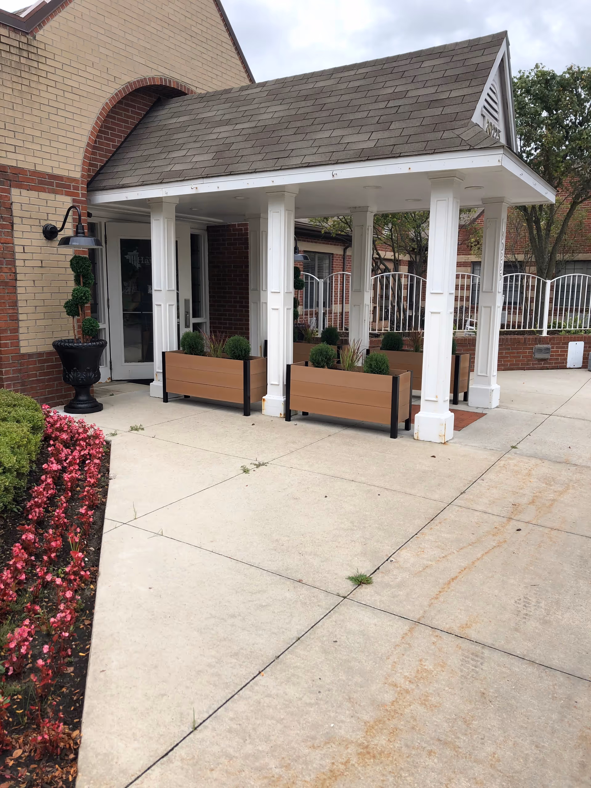 Entrance area of The Haworth Center on the Campus of Baptist Manor featuring a covered porch with white columns, planter boxes with greenery, brick walls, a glass door, and a flower bed with pink flowers along the concrete walkway.