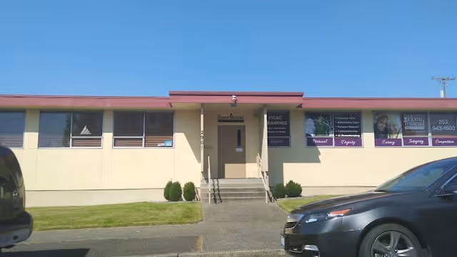 Front exterior view of a single-story building with a beige facade and a red roof trim. There are several windows along the front, a central entrance with stairs and handrails, and a few small bushes near the entrance. Two cars are parked in front of the building on either side of the entrance. Signs with text and images are displayed on the right side of the building's front wall.