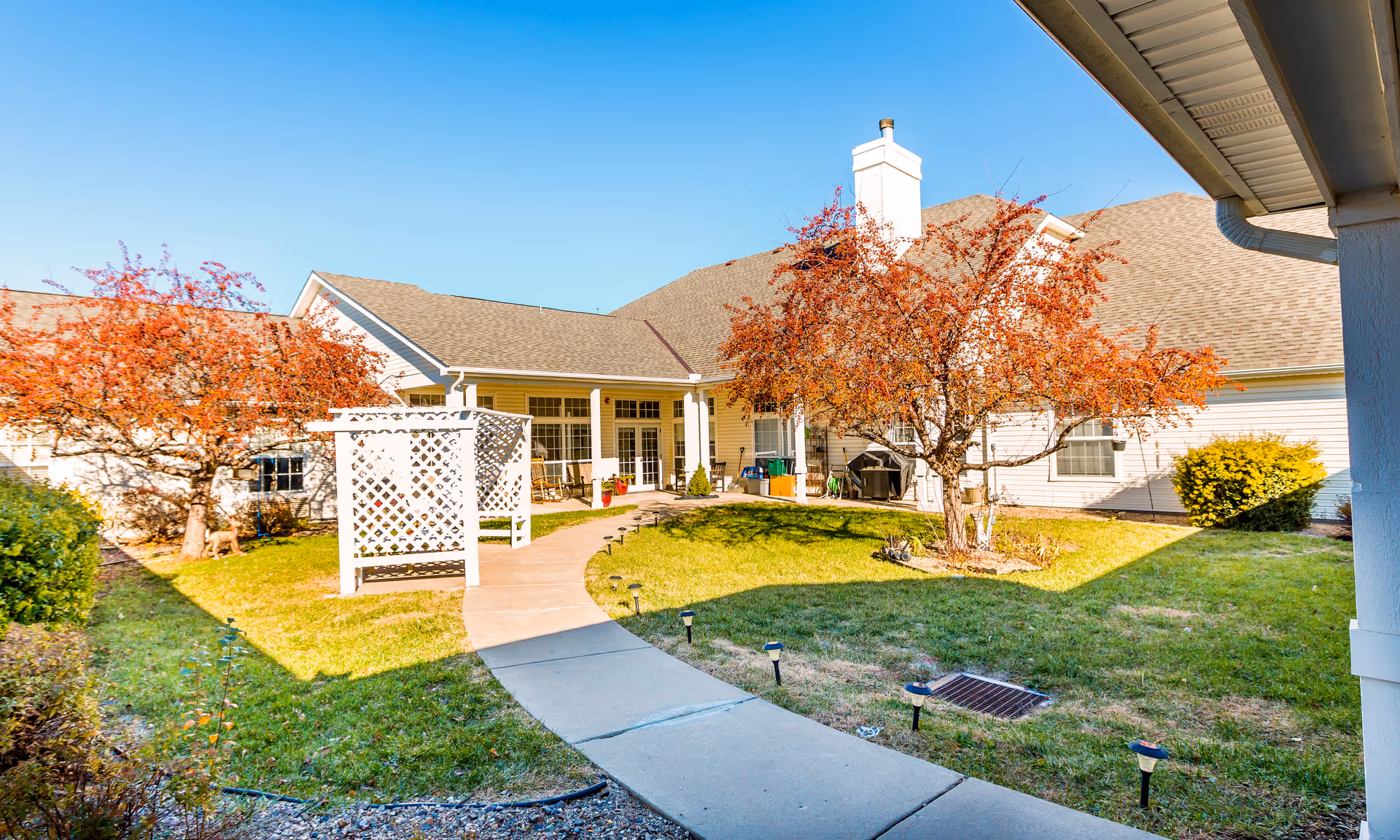 Sunny courtyard with a paved walkway, lattice arbor, autumn trees, and the facility building with a covered patio and seating.