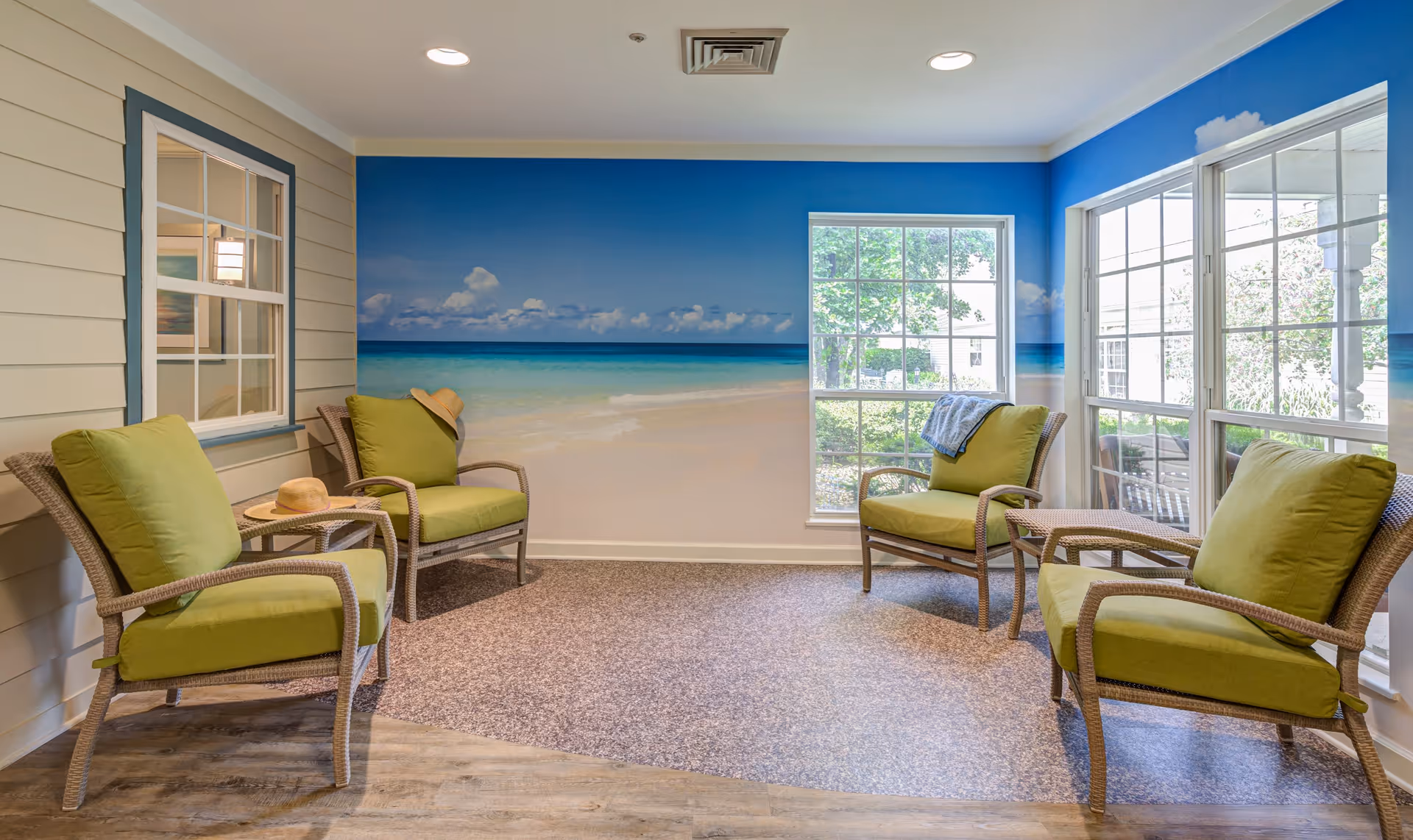 A cozy sitting area with four wicker chairs featuring green cushions arranged in a semi-circle. The walls have a beach mural depicting sand, ocean, and sky, creating a relaxing atmosphere. Large windows allow natural light to brighten the room, and a couple of hats and a blanket are placed on the chairs.