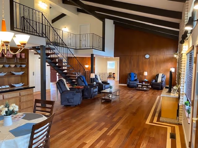Spacious senior living common area with hardwood floors, recliner chairs, a dining table, and a staircase leading to a loft under exposed beams.