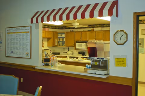 View through a serving window into a kitchen area with wooden cabinets, a microwave, a stove, and beverage dispensers. The serving window has a red and white striped awning above it. A clock and a bulletin board with a calendar are visible on the walls near the window.