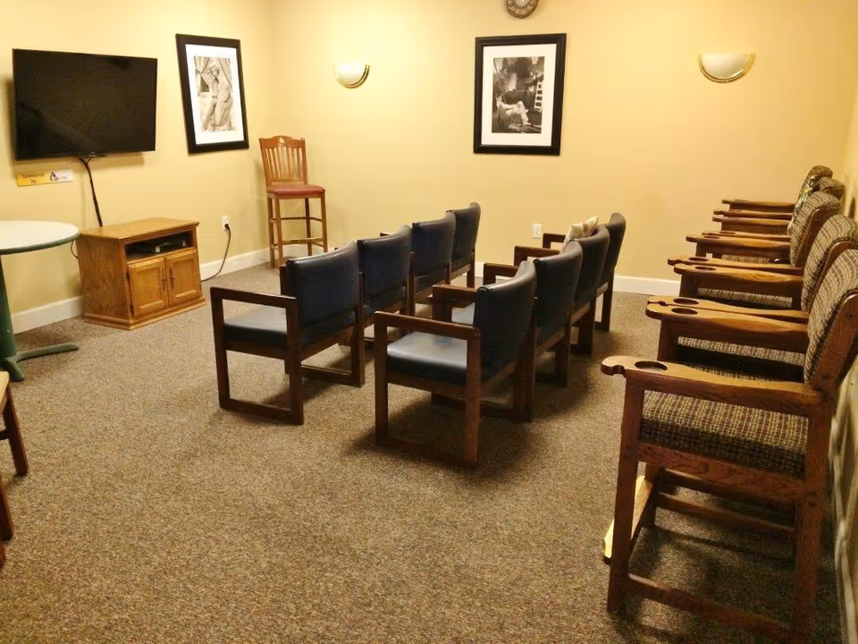 A small room with two rows of chairs facing a wall-mounted flat screen TV. The chairs in the front row have blue cushions and wooden frames, while the chairs in the back row have patterned cushions and wooden armrests with cup holders. There is a wooden cabinet under the TV, a tall wooden chair in the corner, two framed black and white pictures on the wall, and two wall-mounted light fixtures. The room has beige walls and carpeted floor.