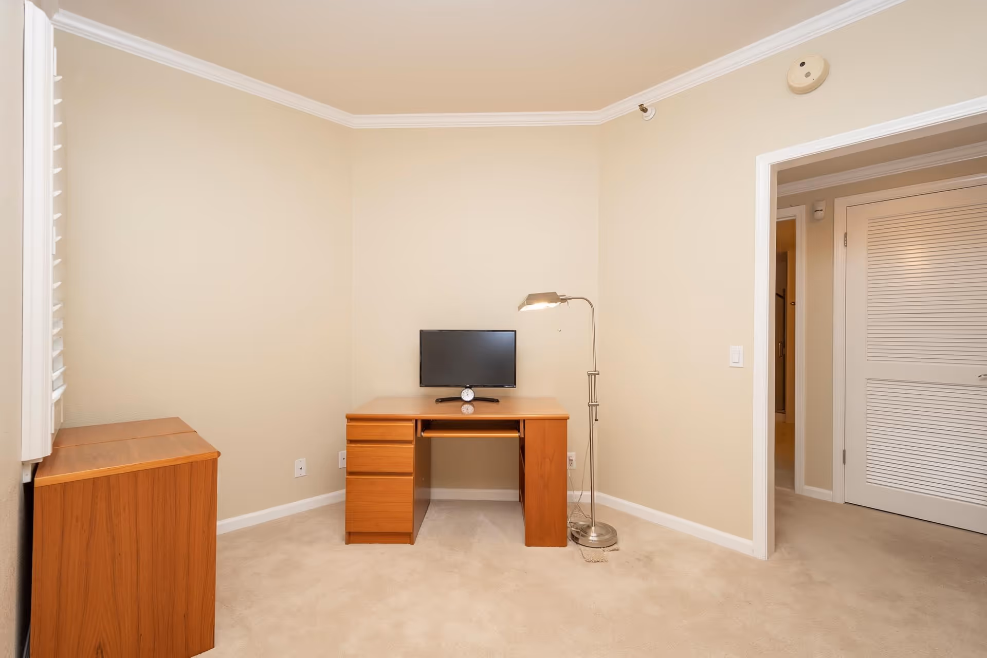Small interior room with a wooden desk holding a monitor, a floor lamp, and beige walls and carpet.