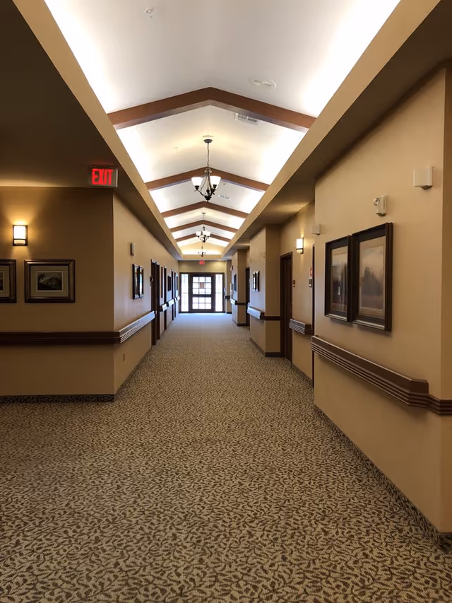 Carpeted interior hallway with vaulted ceiling and chandeliers, handrails, framed artwork and doors leading to rooms.