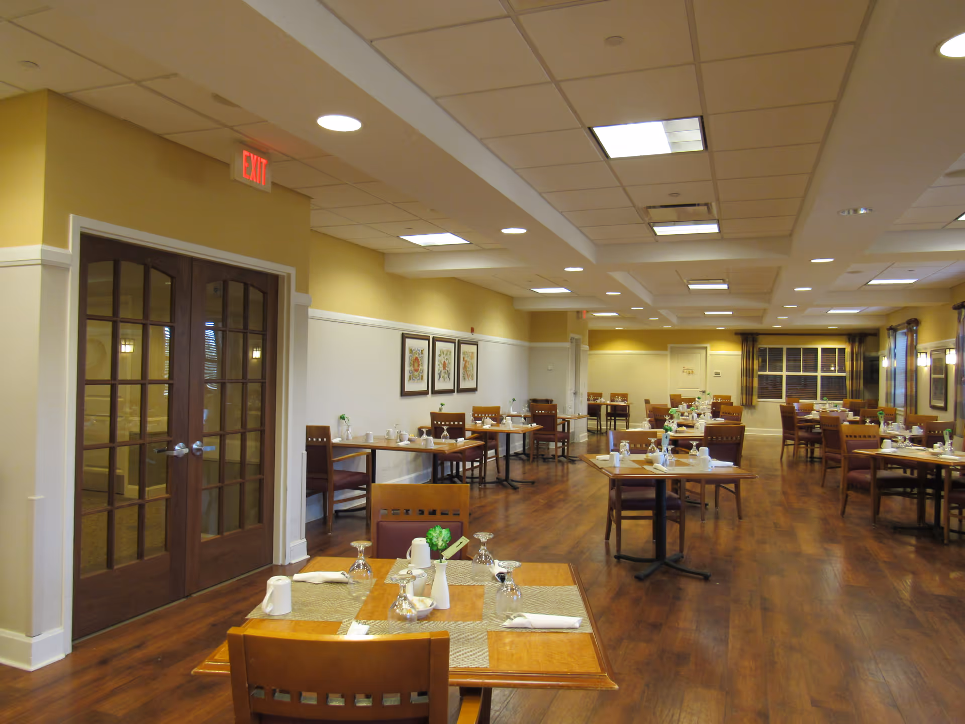 Interior view of a dining room in a senior living facility with wooden tables and chairs arranged neatly. Each table is set with placemats, napkins, glasses, and small flower vases. The room has wooden flooring, light yellow walls with white wainscoting, framed artwork on the walls, and large windows with curtains. There are double wooden doors with glass panels and an exit sign above them.