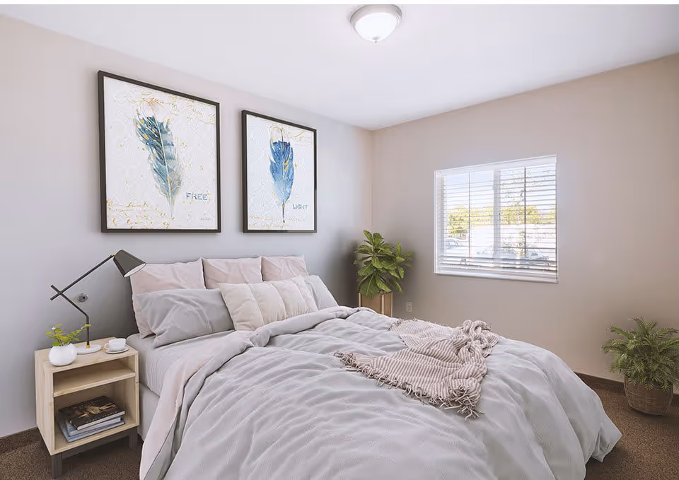 A cozy bedroom with a neatly made bed featuring gray and pink bedding and multiple pillows. Two framed feather artworks hang above the bed, one labeled 'FREE' and the other 'LIGHT'. A small wooden nightstand with a modern lamp, a small plant, and a cup is beside the bed. There is a window with blinds letting in natural light, and two potted plants are placed in the corners of the room.