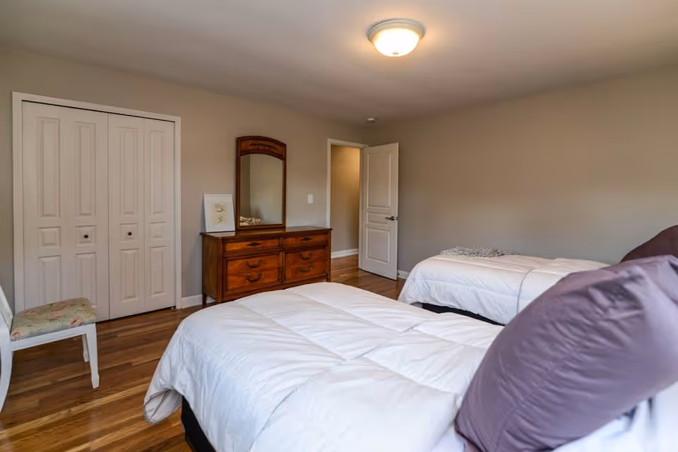 A bedroom with two twin beds covered in white comforters and purple pillows. There is a wooden dresser with a mirror and a framed picture on top against a beige wall. A white chair with a floral cushion is placed near a closed closet with white paneled doors. The room has wooden flooring and a ceiling light fixture.