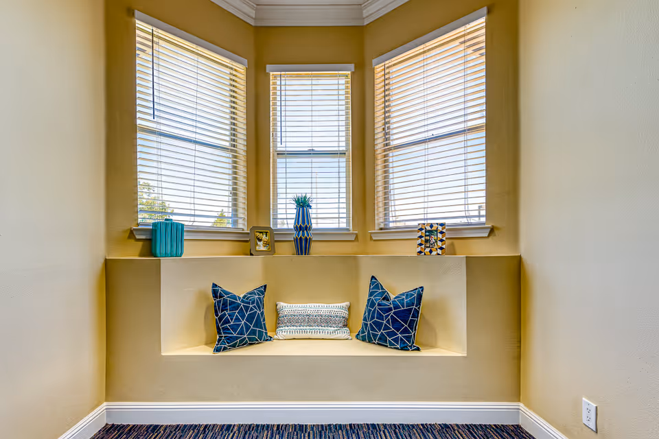 A cozy window nook with three large windows featuring white blinds. The nook has a built-in bench with two blue patterned pillows and one smaller decorative pillow. On the ledge above the bench are three decorative items including a blue vase and two small framed pictures. The walls are painted a warm beige color and the floor is carpeted with a dark striped pattern.