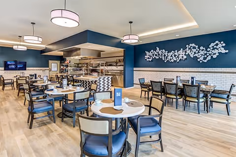 A spacious dining room with multiple round and rectangular tables surrounded by chairs. The room features light wood flooring, blue and white walls, modern ceiling lights, and a decorative wall art piece with white floral designs. An open kitchen area is visible in the background.