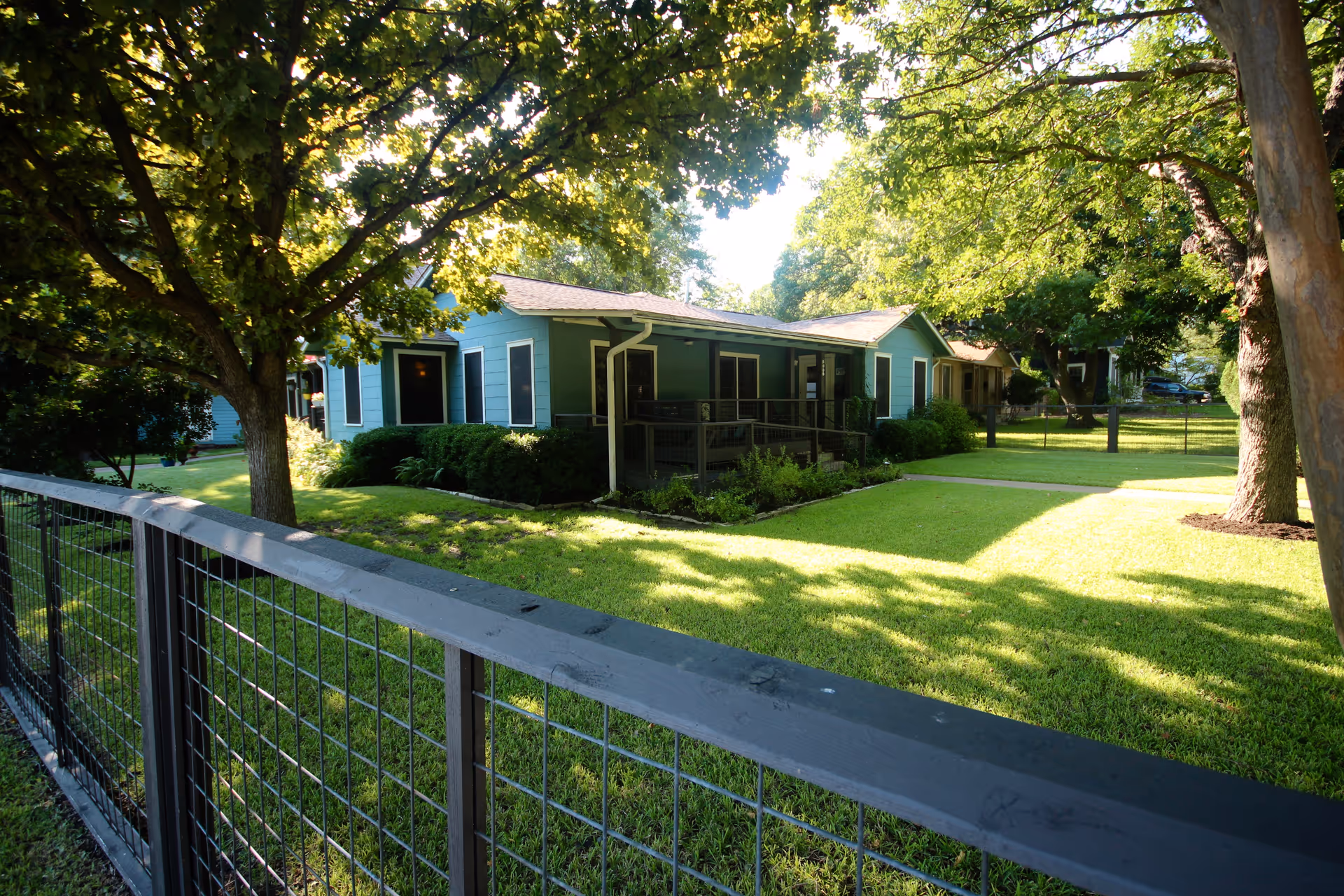 A single-story green building with a porch surrounded by a well-maintained lawn and several large trees. A black metal fence runs along the foreground, enclosing the grassy area. The scene is bright and sunny with shadows cast by the trees.