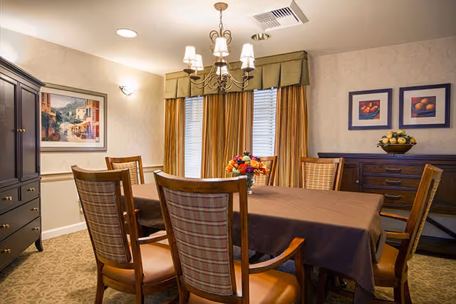 A dining room with a rectangular table covered with a brown tablecloth, surrounded by six wooden chairs with plaid upholstery. A chandelier with six lampshades hangs above the table. The room has patterned carpet, beige walls with framed artwork, and a window with gold curtains and valance. A dark wooden cabinet and sideboard are also visible, with a bowl of fruit on the sideboard.