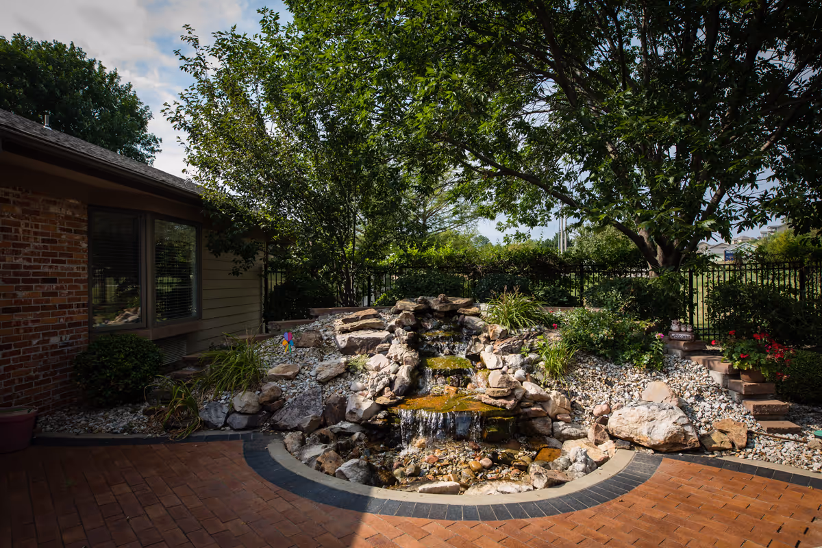Brick patio with a tiered rock waterfall and pond surrounded by landscaping, trees, and the side of a building.