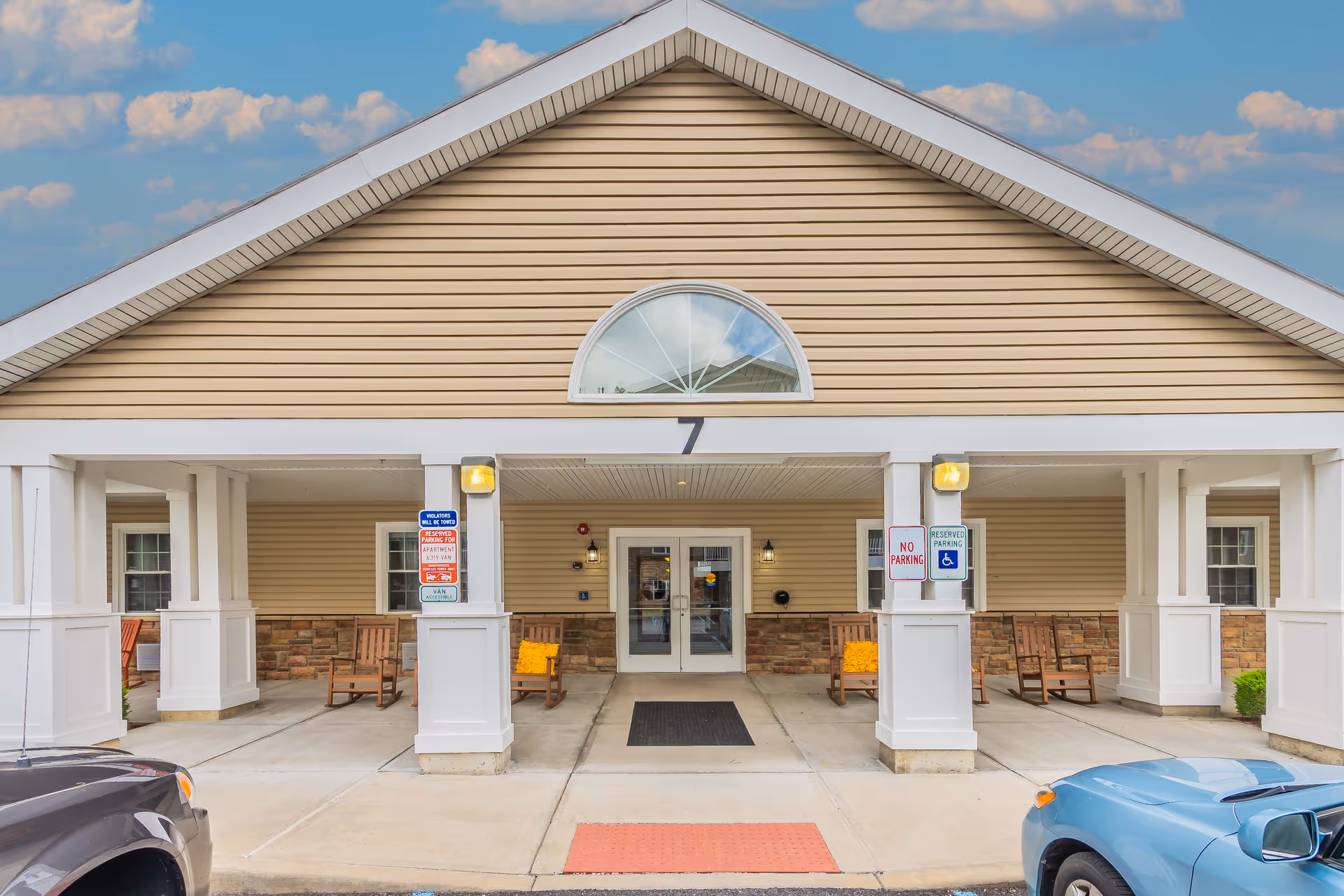 Front entrance of a senior living facility with a covered porch supported by white columns. There are wooden rocking chairs with yellow cushions on the porch. Two cars are partially visible parked in front. The building has beige siding with a stone accent and a large arched window above the entrance doors. Several parking signs are posted on the columns.