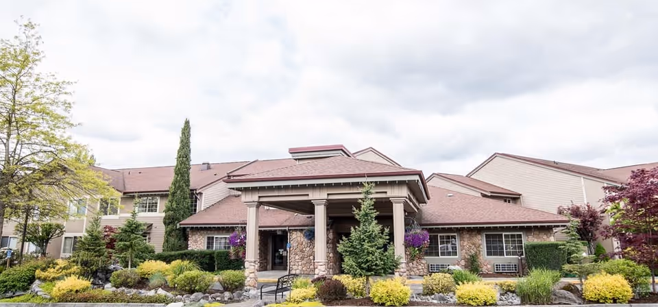 Exterior view of a senior living facility building with a covered entrance supported by stone columns, surrounded by well-maintained landscaping including bushes, trees, and flowers under a cloudy sky.