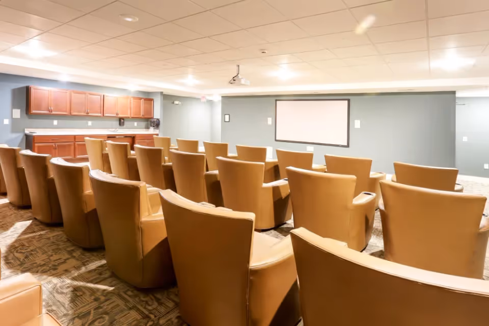 A small screening/meeting room with rows of tan leather chairs facing a projector screen and cabinets along the side.