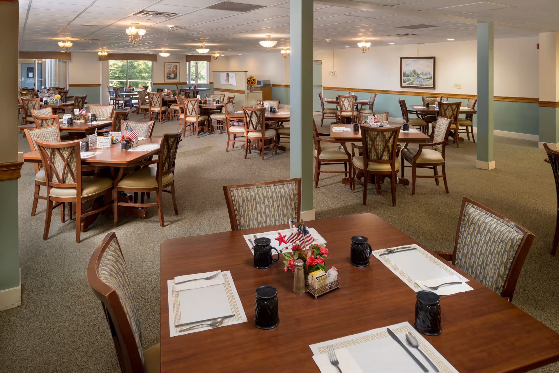 A spacious dining room with multiple wooden tables and chairs arranged neatly. Each table is set with placemats, utensils, black cups, and small centerpieces featuring flowers and miniature American flags. The room has carpeted floors, light green and white walls with wooden trim, and several ceiling light fixtures. Large windows allow natural light to brighten the space, and framed artwork hangs on the walls.