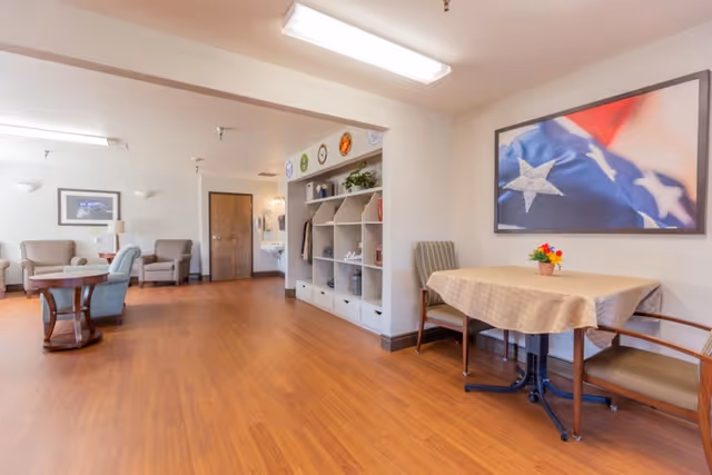 A spacious, well-lit common area with wooden flooring featuring a small table with two chairs and a vase of flowers, a large framed picture of an American flag on the wall, a built-in shelving unit with decorative items, and several armchairs arranged around a round wooden table in the background.