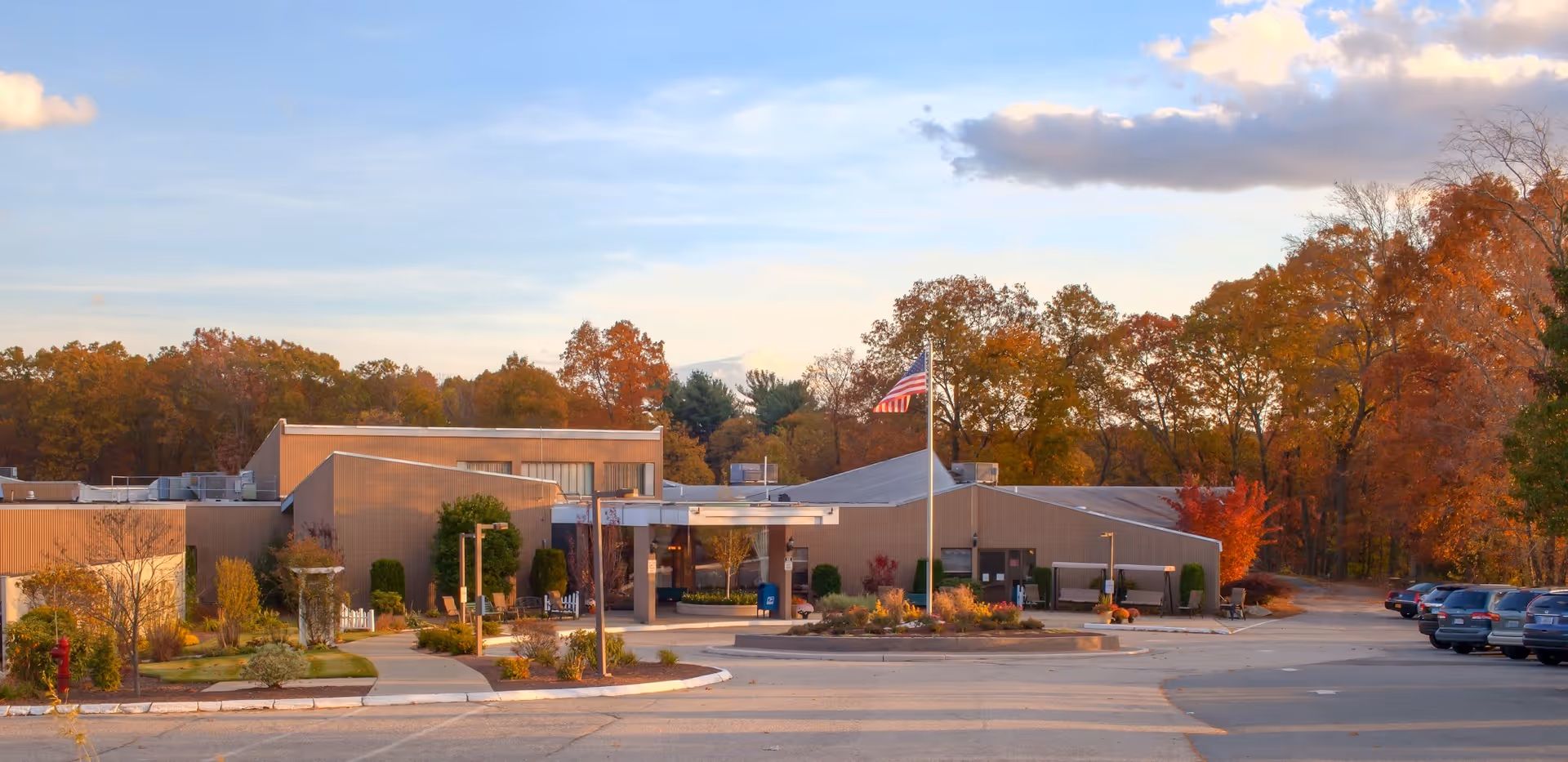 Single-story senior living facility building with an American flag at the entrance, surrounded by autumn trees and a parking lot.