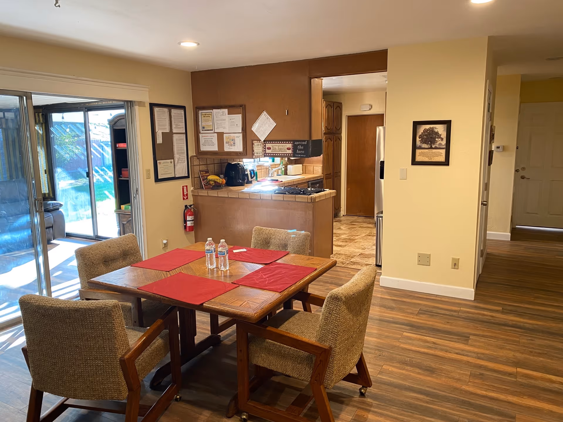 Interior view of a dining area in an elderly care facility with a wooden table set with four cushioned chairs, red placemats, and two water bottles. The room has wood flooring and opens into a kitchen with tiled countertops and cabinets. A sliding glass door leads to an outdoor space with visible greenery.
