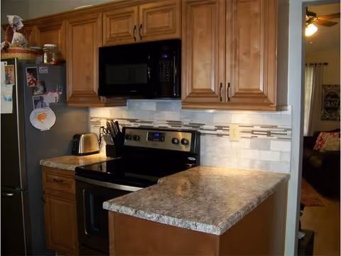 A kitchen area with wooden cabinets, a black microwave mounted above a black electric stove, a toaster on the counter, and a refrigerator with magnets and photos. The countertop is a light-colored granite or laminate, and there is a tiled backsplash with a decorative horizontal strip. In the background, a living room area with a ceiling fan and a wall decoration is partially visible.