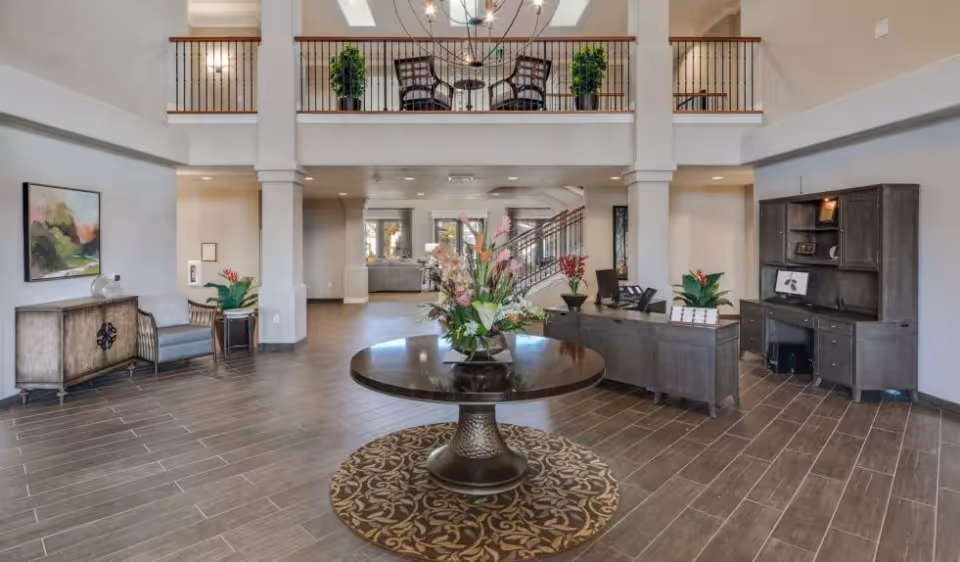 Spacious and well-lit lobby area with a round table featuring a large floral arrangement in the center. The room has a high ceiling with a balcony railing above, two armchairs and a sideboard on the left, and a reception desk with a hutch on the right. The floor is tiled with a wood-like pattern, and there are decorative plants placed around the space.
