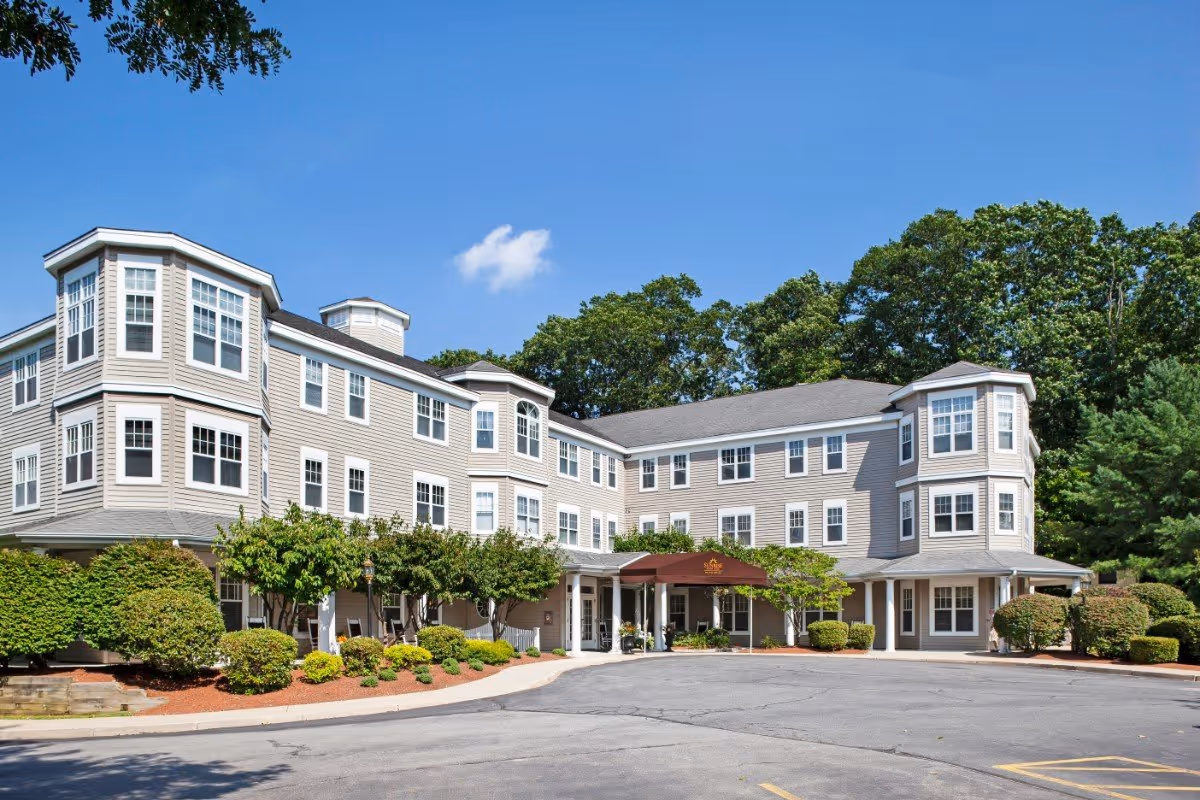 Three-story beige senior living facility building with a covered entrance, landscaped shrubs, and a paved driveway under a clear blue sky.