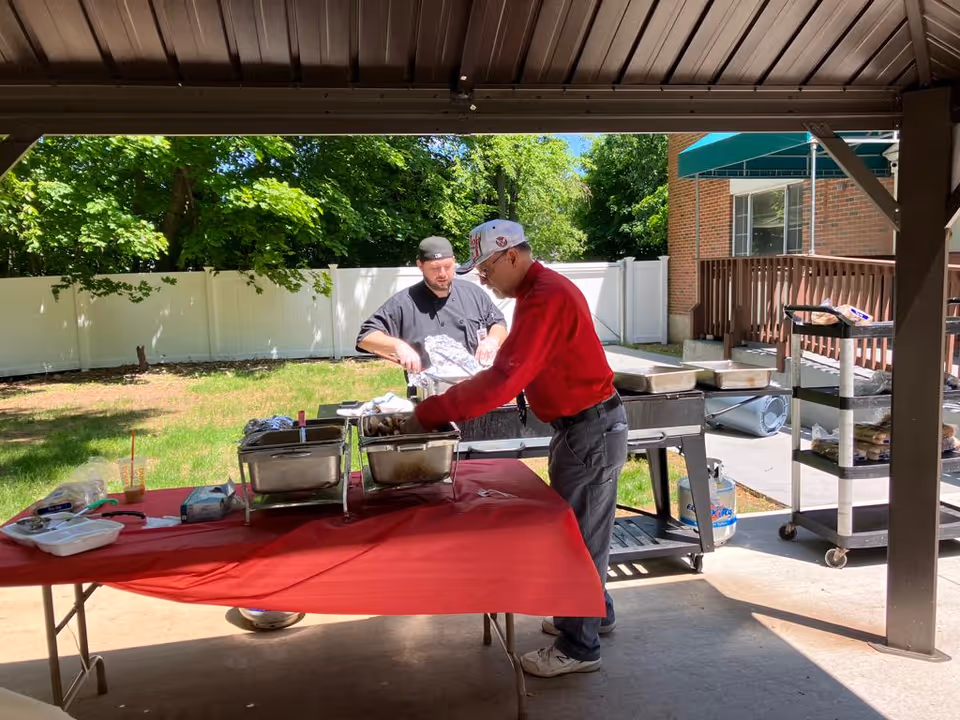 Two men preparing food outdoors under a covered area. One man in a black chef uniform stands behind a table with food trays, while another man in a red shirt and white cap serves food from the trays. There is a grassy area and trees in the background, along with a white fence and part of a brick building with a green awning.