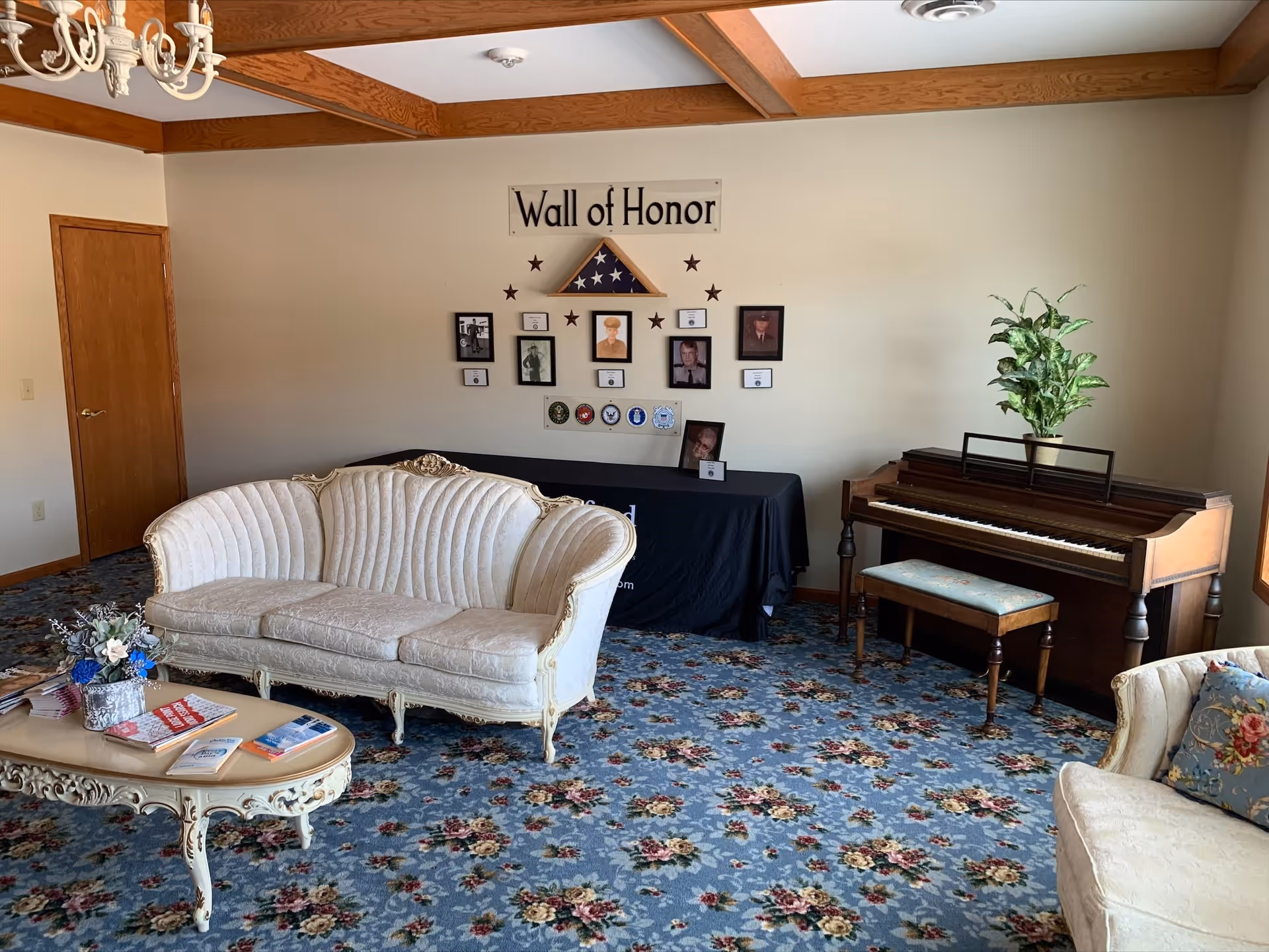 A senior living facility common area with vintage-style white upholstered sofas, a floral carpet, a wooden piano with a plant on top, and a 'Wall of Honor' display featuring framed photos, a folded American flag, and military insignias on the wall.