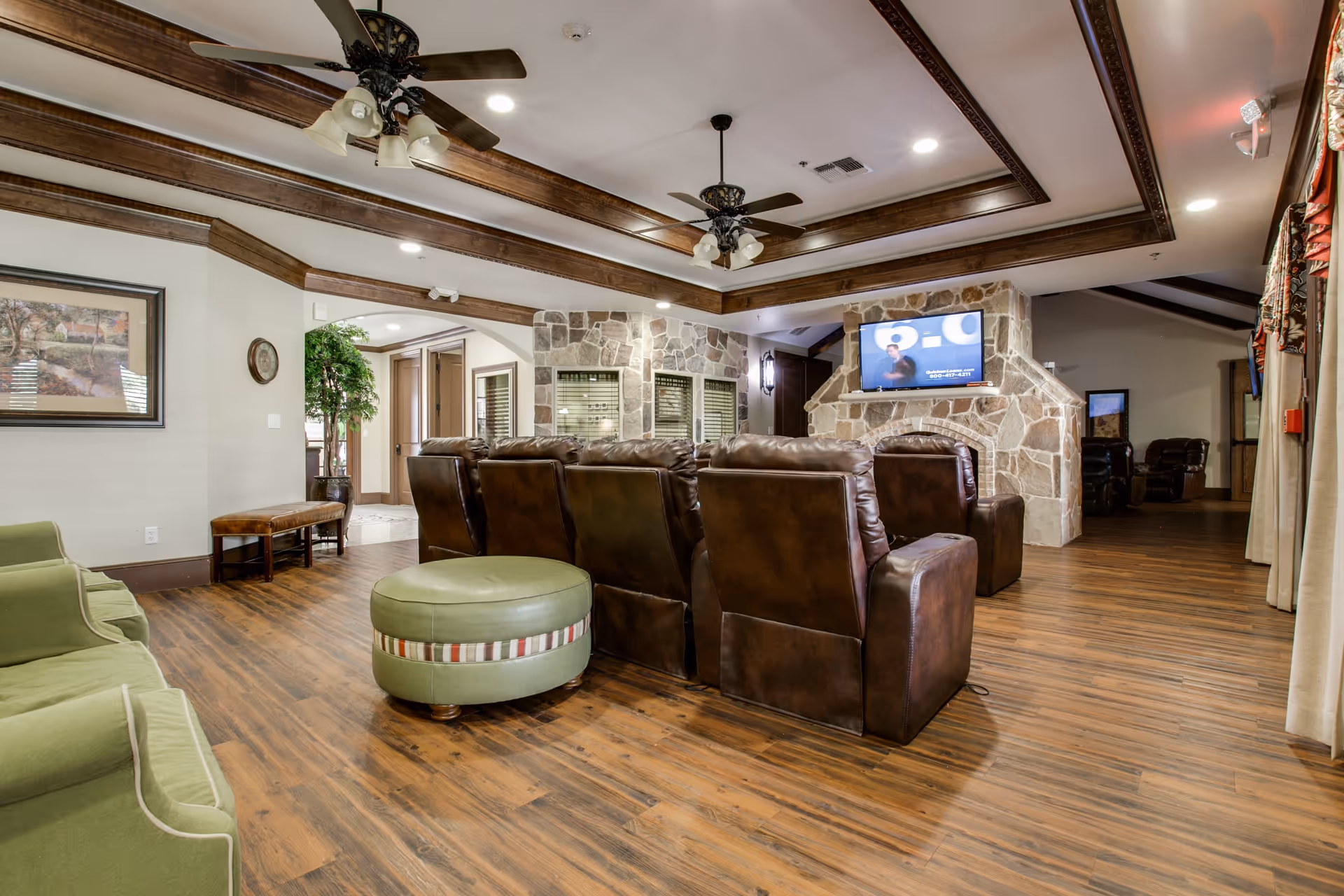 A spacious living room area with wooden flooring, a stone fireplace with a mounted flat-screen TV, and multiple brown leather recliners arranged facing the TV. The ceiling features wooden beams and two ceiling fans with lights. There are green upholstered chairs and a round green ottoman with a striped band in the foreground. The room has neutral-colored walls with framed artwork and a large window with curtains on the right side.
