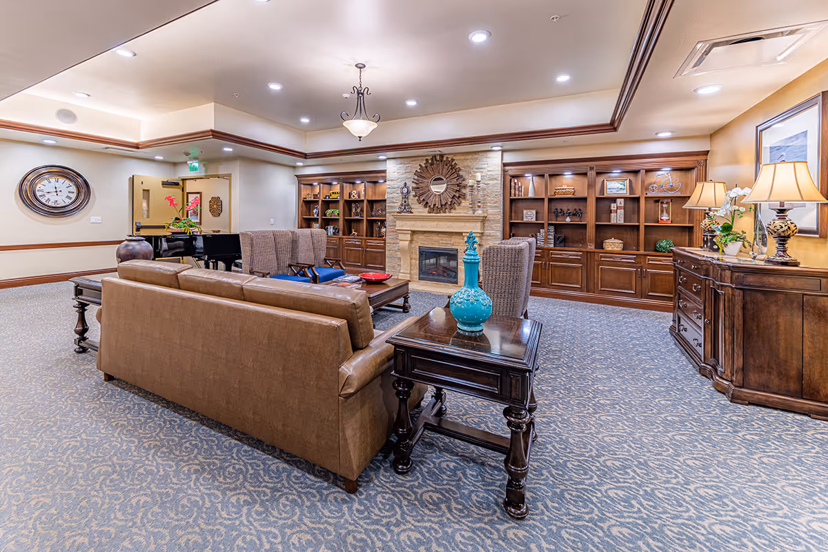 A cozy senior living room with a brown leather sofa, two upholstered armchairs, a wooden coffee table, and a side table with a turquoise decorative vase. The room features built-in wooden bookshelves filled with decorative items, a stone fireplace with a sunburst mirror above it, and warm lighting from ceiling fixtures and table lamps. The carpet has a blue and beige pattern, and there is a large wall clock and a piano in the background.