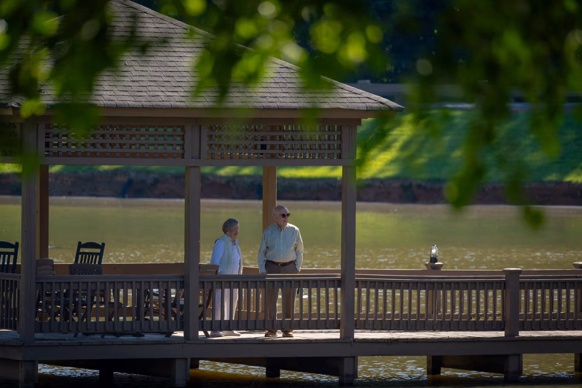An elderly man and woman standing together on a wooden gazebo overlooking a calm lake, surrounded by greenery and shaded by tree branches.