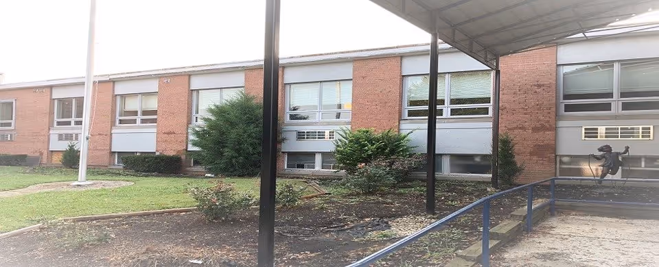 Front exterior of a single-story brick building with rows of windows, shrubs, and a covered walkway/entrance.