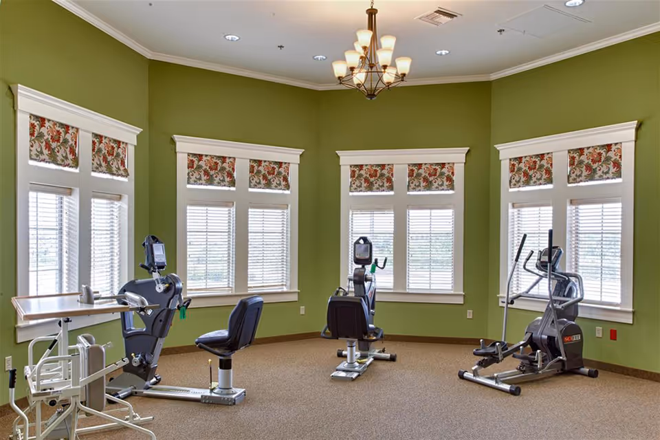 A fitness room with green walls and four large windows with floral valances. The room contains three exercise machines including two recumbent bikes and one elliptical machine. There is also a small table with a chair on wheels. The floor is carpeted and a chandelier hangs from the ceiling.