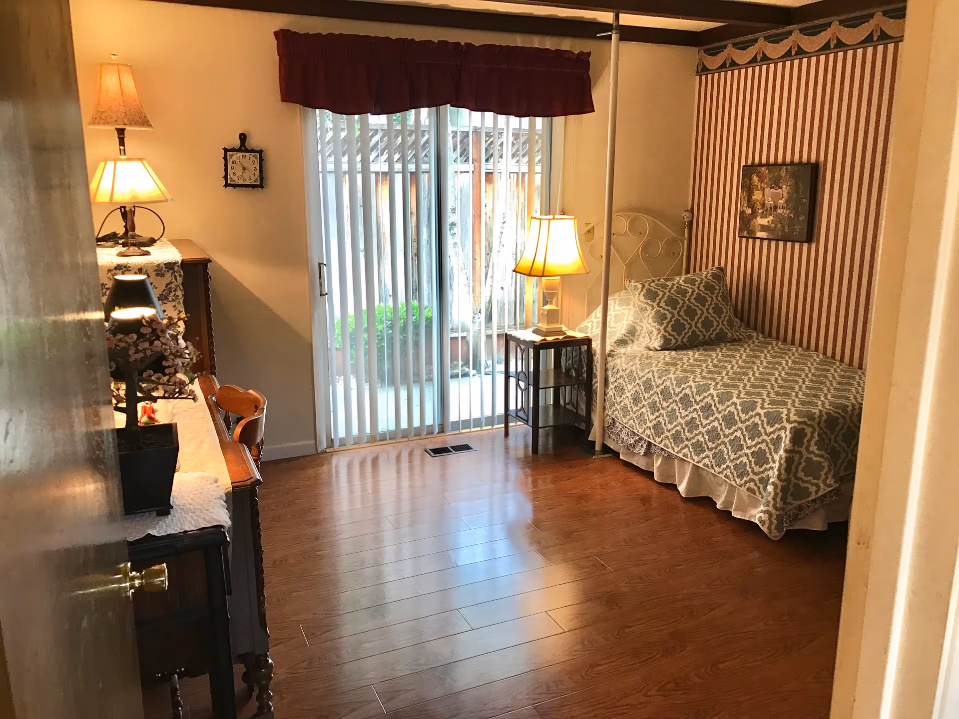 A cozy bedroom with a single bed covered in a patterned blue and white bedspread and matching pillow. There is a small bedside table with a lamp next to the bed. The room has wooden flooring and a sliding glass door with vertical blinds leading to an outdoor area. On the left side, there is a wooden dresser with a lamp and decorative items on top. The walls are decorated with a framed picture and striped wallpaper behind the bed.