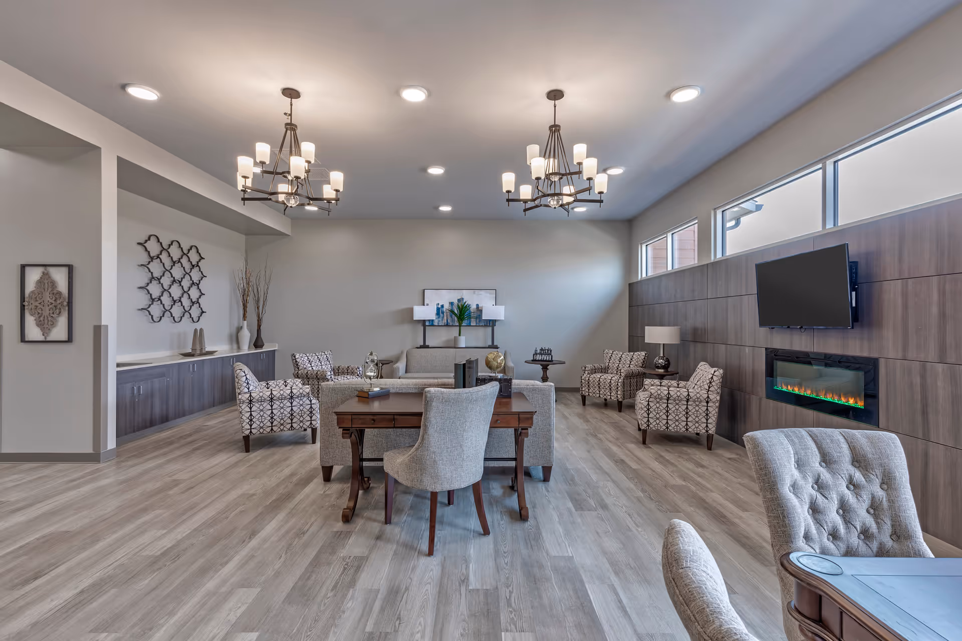 A spacious, modern living room with light gray wood flooring, multiple upholstered chairs and sofas arranged around a wooden table, two chandeliers hanging from the ceiling, a wall-mounted flat-screen TV above an electric fireplace, and decorative wall art and lamps.