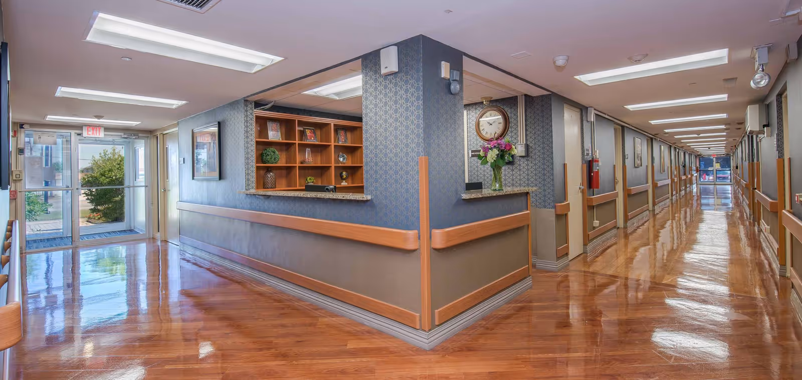 A long, polished wooden floor hallway in a senior living facility with handrails on both sides. To the left, there is a reception desk with shelves holding decorative items and a clock on the wall above a vase of flowers. The hallway has multiple doors along the right side and large glass exit doors at both ends, allowing natural light to enter.