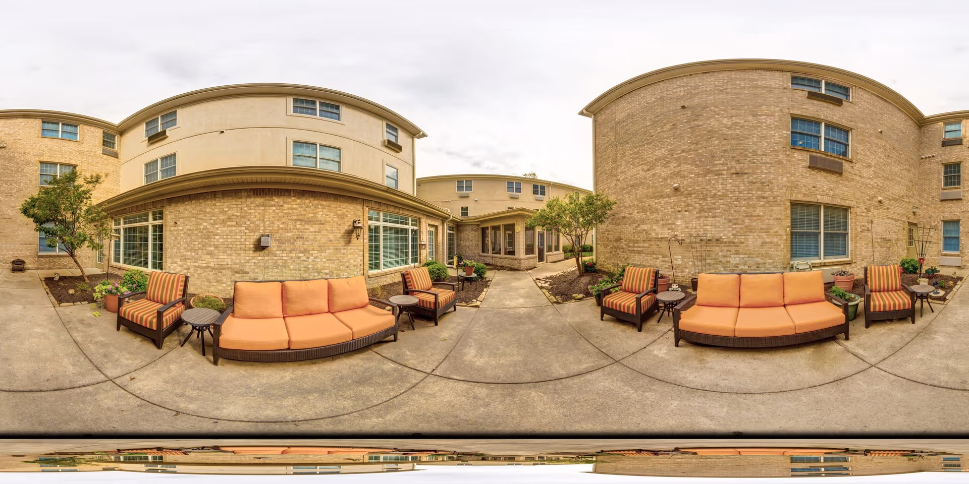 Outdoor courtyard area of a senior living facility with beige brick buildings surrounding a concrete patio. The patio features orange cushioned sofas and striped cushioned armchairs with small round tables. There are potted plants and small trees along the edges of the patio.