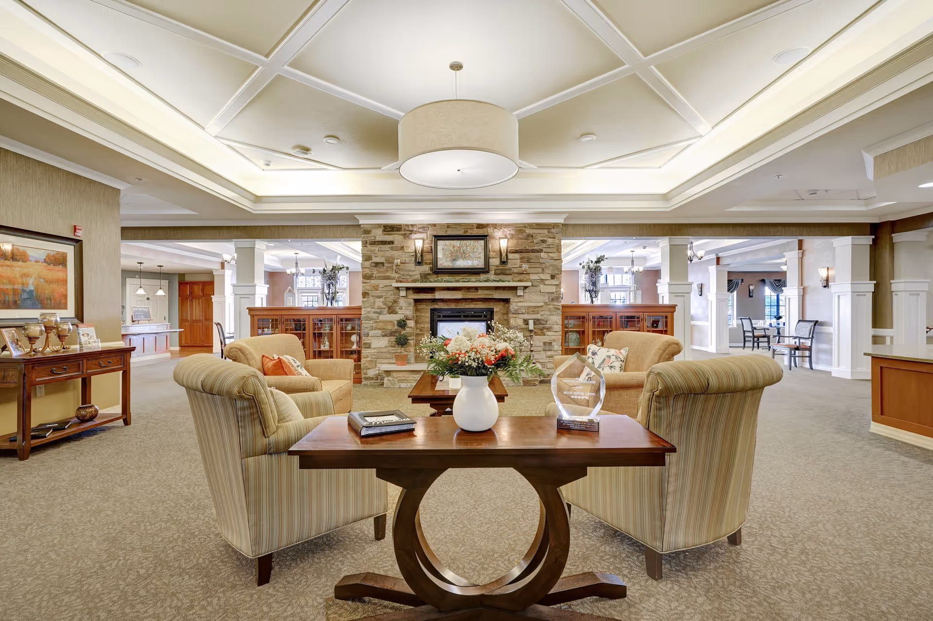 A spacious and well-lit senior living common area featuring a stone fireplace in the center, surrounded by comfortable armchairs and a wooden coffee table with a white vase of flowers. The room has a coffered ceiling with recessed lighting, beige carpeting, and decorative columns. In the background, there are additional seating areas and large windows letting in natural light.