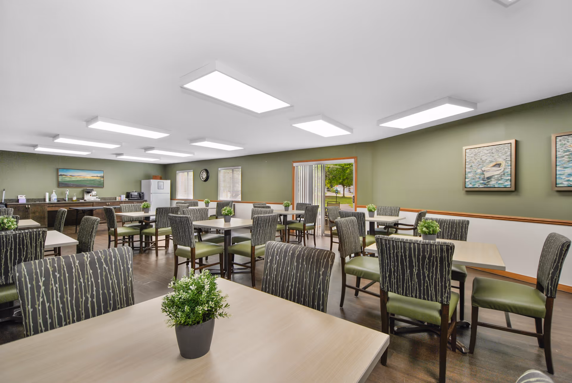 A dining room with multiple tables and chairs arranged neatly. Each table has a small green potted plant as decoration. The walls are painted green with two paintings of boats on water. There are large rectangular ceiling lights providing bright illumination. A refrigerator, microwave, and coffee machine are visible along the back wall. A sliding glass door opens to an outdoor area with greenery visible outside.
