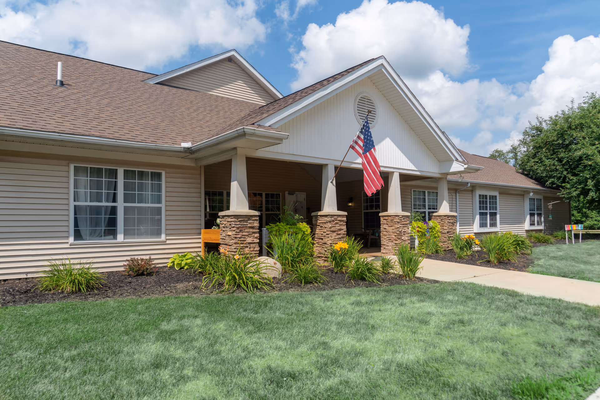 Front entrance of a single-story senior living building with a covered porch, American flag, and landscaped lawn.