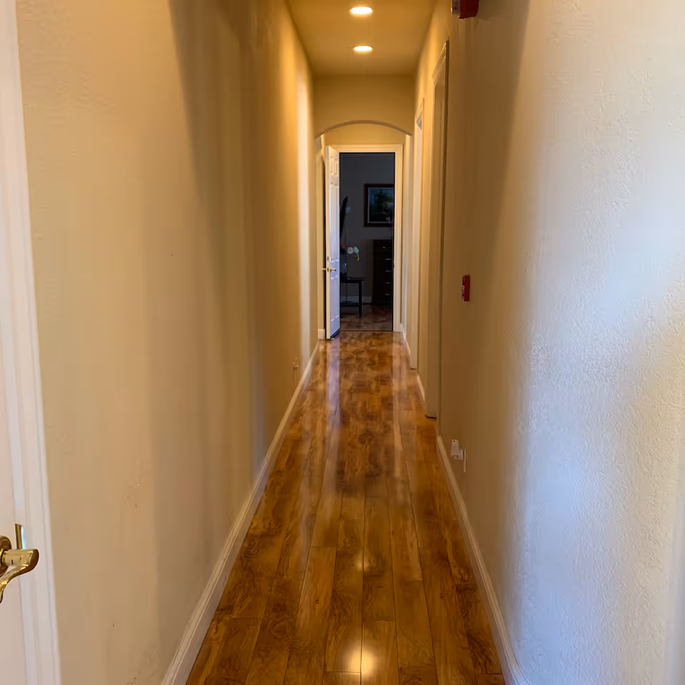 Long narrow interior hallway with glossy wood flooring, cream walls, recessed ceiling lights and an open doorway at the far end.