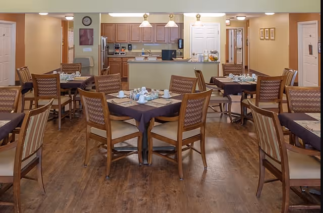 A dining room in a senior living facility with multiple wooden tables and chairs arranged neatly. Each table is covered with a dark tablecloth and set with cups, saucers, and utensils. In the background, there is a kitchen area with wooden cabinets, a countertop, and various kitchen appliances. The room has warm lighting and a wooden floor.