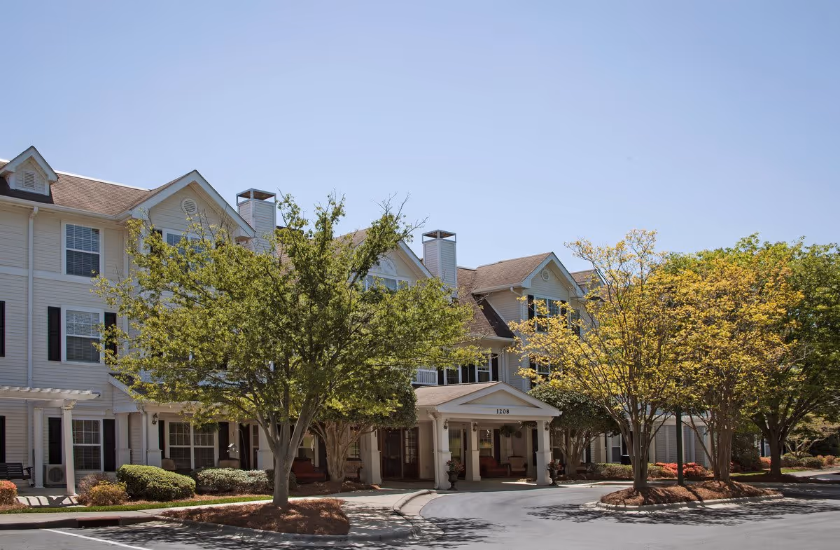 Exterior view of a multi-story senior living facility building with a covered entrance, surrounded by trees and landscaping under a clear blue sky.