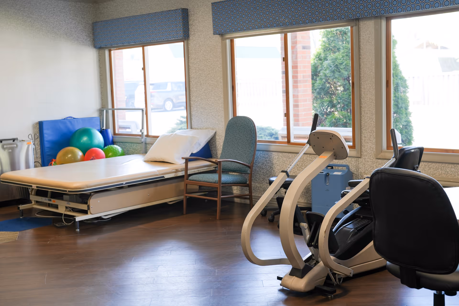 A room with exercise equipment including a recumbent bike, a padded therapy table with a pillow, a chair, and colorful exercise balls near the windows with blue valances. The room has wooden flooring and large windows letting in natural light.